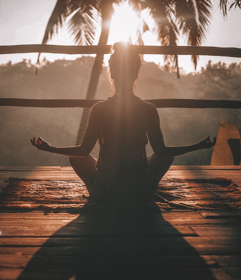 Person meditating in lotus pose on a wooden deck at sunset with palm trees